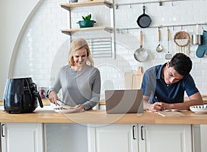 Smiling beautiful mature woman cooking toast bread by Air Fryer machine for breakfast at kitchen table while husband using laptop