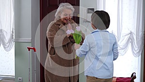 Smiling beautiful mature woman standing with travel bag as teen boy walking with bouquet of flowers hugging. Live camera
