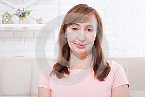 Smiling Beautiful middle-aged brunette woman sitting on sofa at home looking at the camera. Face with wrinkles. Menopause.