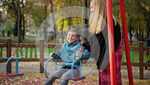 Smiling beautiful mother swinging son on swings outdoors in autumn park. Portrait of happy relaxed Caucasian woman