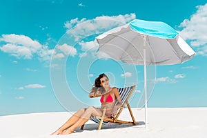 Beautiful sexy girl in swimsuit and sunglasses sitting in deck chair under umbrella on sandy beach
