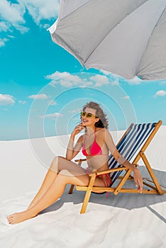 Beautiful sexy girl in swimsuit and sunglasses sitting in deck chair under umbrella on sandy beach