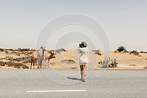 A smiling beautiful tourist girl takes pictures of a camel by the road while traveling. Vacation, trip, vacation