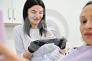 Smiling beautiful woman dentist orthodontist holding teeth color and shade chart guide according to Vita scale.