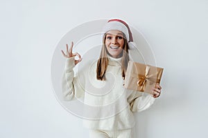 Smiling beautiful woman in red santa claus outfit holding gift and showing okay gesture isolated on the white background