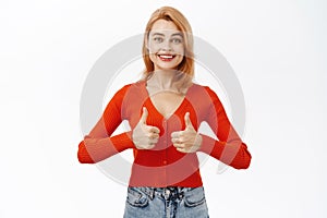 Smiling beautiful woman shows thumbs up in approval. Girl likes and approves smth, stands over white background