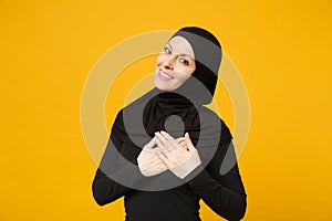 Smiling beautiful young arabian muslim woman in hijab black clothes put hands on heart isolated on yellow wall