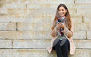 Smiling beautiful young woman sitting on stairs using mobile phone on winter day