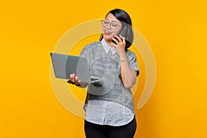 Smiling beautiful young woman talking on smartphone and looking at laptop screen on yellow background