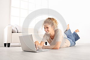 smiling Beautiful young woman using a laptop, lying on living room wooden floor in modern white home