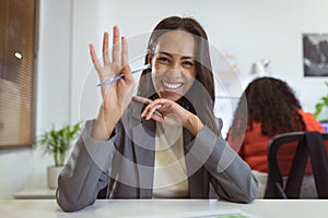 Smiling biracial businesswoman sitting at desk making video call waving in modern office