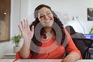 Smiling biracial businesswoman sitting at desk making video call waving in modern office