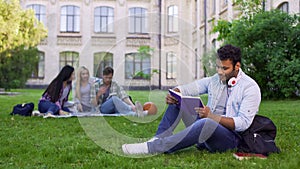 Smiling biracial male student sitting on grass and reading interesting book