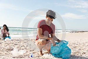 Smiling biracial man collecting garbage in blue plastic bag from sand at beach against sky