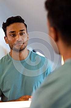 Smiling biracial man looking at himself in bathroom mirror after applying face cream