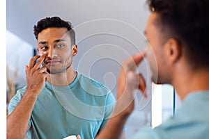 Smiling biracial man looking at himself in bathroom mirror and applying face cream