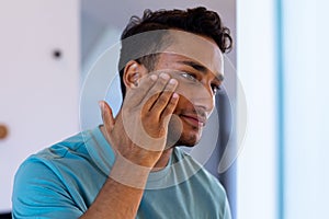 Smiling biracial man looking at himself in bathroom mirror and applying face cream