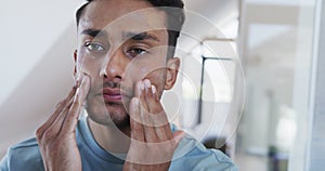 Smiling biracial man looking in mirror and applying face cream in bathroom, slow motion