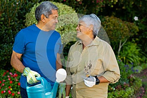 Smiling biracial senior couple holding watering can and gardening fork looking at each other in yard