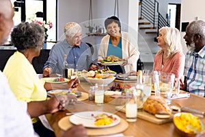 Smiling biracial senior woman serving food to multiracial friends at dining table in nursing home