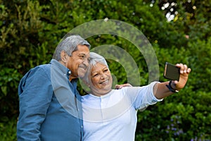 Smiling biracial senior woman taking selfie with husband over smart phone against trees in park