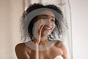 Smiling biracial woman apply under eye cream looking in mirror