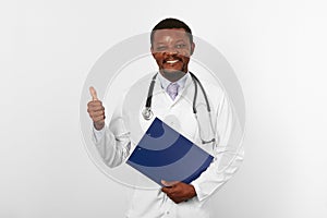 Smiling black bearded doctor man in white robe holds medical clipboard and shows thumbs up gesture