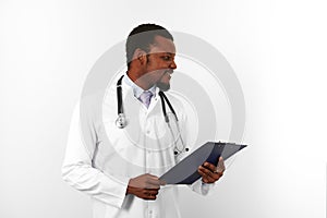 Smiling black bearded doctor man in white robe with stethoscope holds medical chart on clipboard