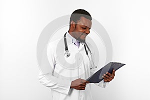 Smiling black bearded doctor man in white robe with stethoscope looks to medical chart on clipboard