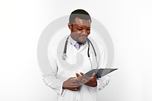 Smiling black bearded doctor man in white robe with stethoscope looks to medical chart on clipboard
