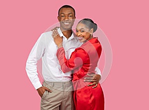 Smiling black couple embracing, woman in red dress, man in white shirt, pink background