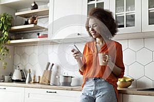 Smiling Black Female Messaging On Smartphone And Drinking Coffee In Kitchen Interior