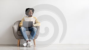 Smiling black guy with laptop computer sitting in chair and looking aside