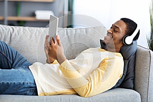 Smiling black guy laying on couch, watching movies on pad