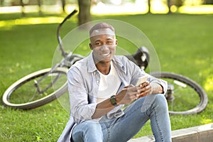 Smiling black guy with mobile phone sitting near his bicycle at park