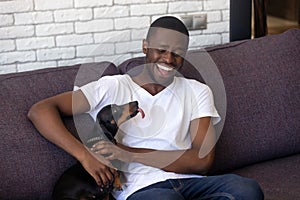 Smiling black guy relax on sofa playing with dackel dog photo