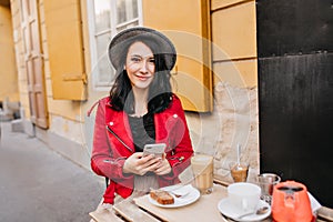 Smiling black-haired girl with phone sitting in outdoor cafe in morning. Photo of cheerful lady in hat and red jacket