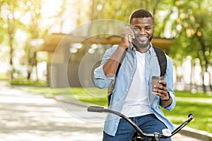 Smiling black man on bicycle holding coffee and talking on smartphone