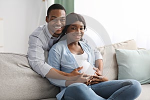 Smiling black man hugging his pregnant woman sitting on sofa