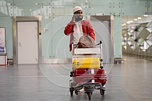 Smiling Black man pushing luggage trolley walking after arrival at airport, talking on mobile phone.