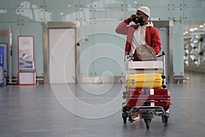 Smiling Black man pushing luggage trolley walking after arrival at airport, talking on mobile phone.