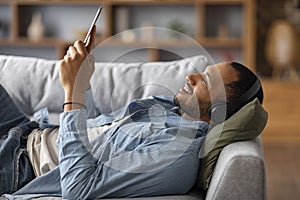 Smiling Black Man Wearing Wireless Headphones Resting With Digital Tablet At Home