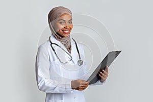 Smiling black muslim female doctor in hijab with clipboard in hands