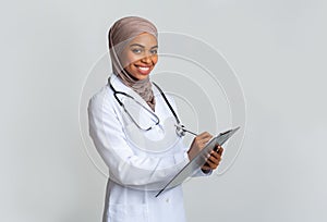 Smiling black muslim female doctor in hijab writing notes to clipboard