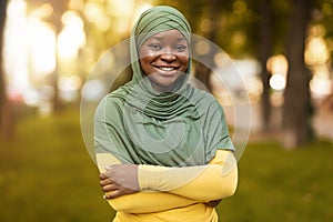 Smiling Black Muslim Woman In Hijab Standing With Folded Arms Outdoors