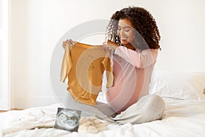 Smiling black pregnant woman demonstrating charming mustard-colored baby onesie,