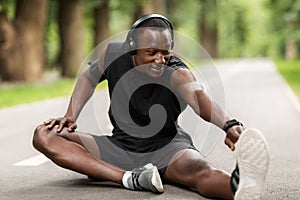 Smiling black sportsman sitting on park path and stretching