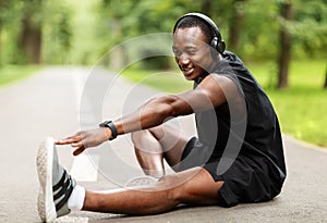 Smiling black sportsman sitting on pathway at park, stretching