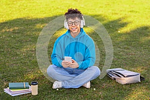Smiling black student guy wearing headphones sitting with phone outside