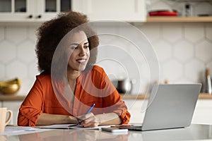 Smiling Black Student Woman Using Laptop In Kitchen And Taking Notes